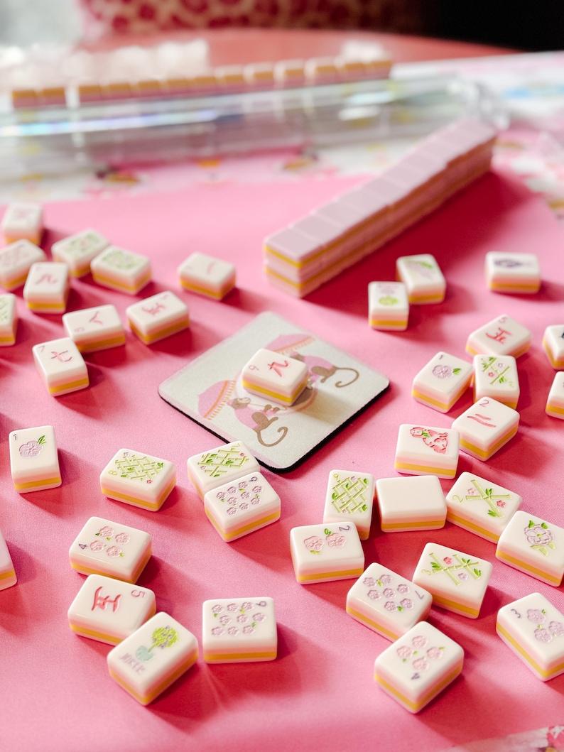 May include: A collection of Mahjong tiles with a pink and yellow color scheme, scattered on a pink surface. The tiles feature various symbols and floral designs. A matching tile rack and a coaster with a monkey design are also present.