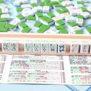 May include: A Mahjong set with white tiles featuring green and blue designs, numbers, and letters. The tiles are arranged on a wooden rack, with additional tiles scattered around. A game guide is visible below.