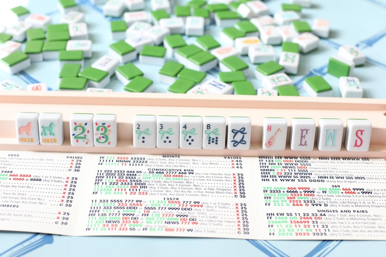 May include: A Mahjong set with white tiles featuring green and blue designs, numbers, and letters. The tiles are arranged on a wooden rack, with additional tiles scattered around. A game guide is visible below.