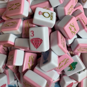 May include: A collection of pink and white Mahjong tiles. The tiles feature various symbols, numbers, and characters in gold and pink. Some tiles display a diamond, a ring, and the word "NORTH".