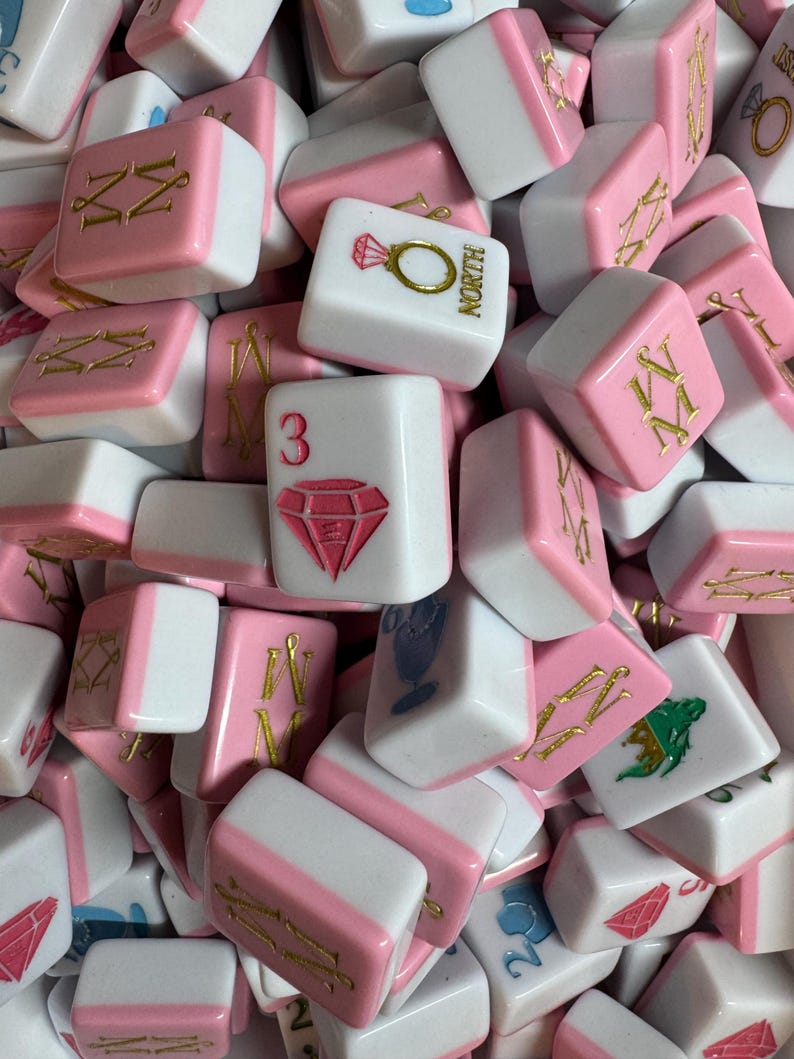 May include: A collection of pink and white Mahjong tiles. The tiles feature various symbols, numbers, and characters in gold and pink. Some tiles display a diamond, a ring, and the word "NORTH".