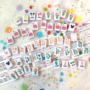 May include: A collection of small, square tiles with various designs, including flowers, purses, and the word "Joker." The tiles are scattered on a white surface with colorful confetti and gold glitter. Some tiles have numbers and directional symbols.