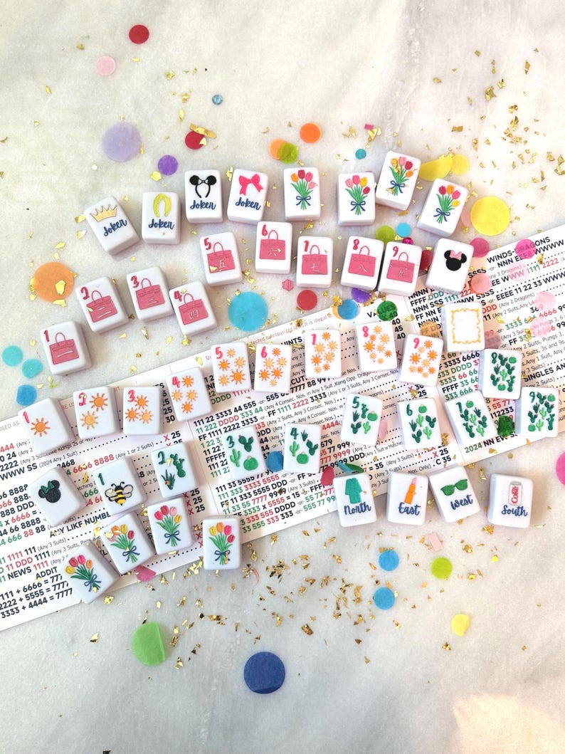 May include: A collection of small, square tiles with various designs, including flowers, purses, and the word "Joker." The tiles are scattered on a white surface with colorful confetti and gold glitter. Some tiles have numbers and directional symbols.