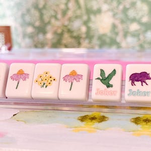 May include: Six white Mahjong tiles with floral and animal illustrations. The tiles feature images of flowers, a bird, and a boar. Two tiles have the word "Joker" printed on them in a teal color.