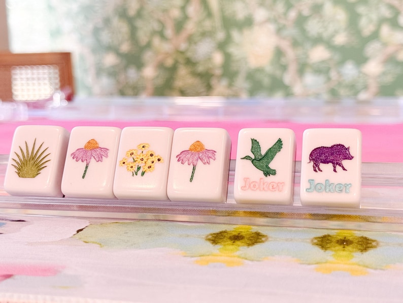 May include: Six white Mahjong tiles with floral and animal illustrations. The tiles feature images of flowers, a bird, and a boar. Two tiles have the word "Joker" printed on them in a teal color.
