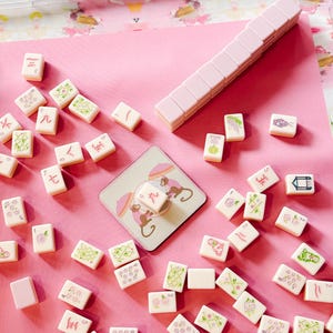 May include: A pink and white Mahjong set with tiles scattered on a pink surface. The tiles feature various symbols and illustrations in green and pink. A stack of pink tiles and a coaster with monkey illustrations are also visible.