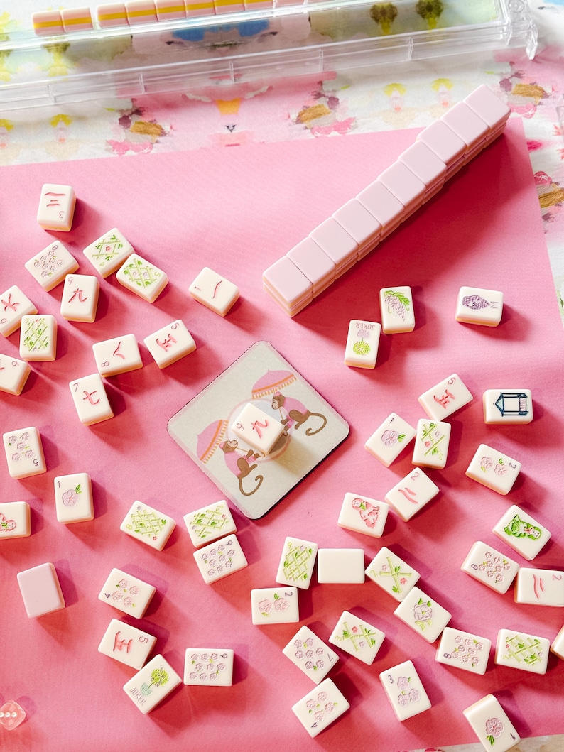 May include: A pink and white Mahjong set with tiles scattered on a pink surface. The tiles feature various symbols and illustrations in green and pink. A stack of pink tiles and a coaster with monkey illustrations are also visible.