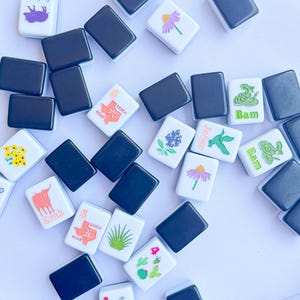 May include: A collection of white and black Mahjong tiles scattered on a white surface. The white tiles feature various colorful illustrations, including flowers, a longhorn, a cactus, and the text "Joker" and "Bam".