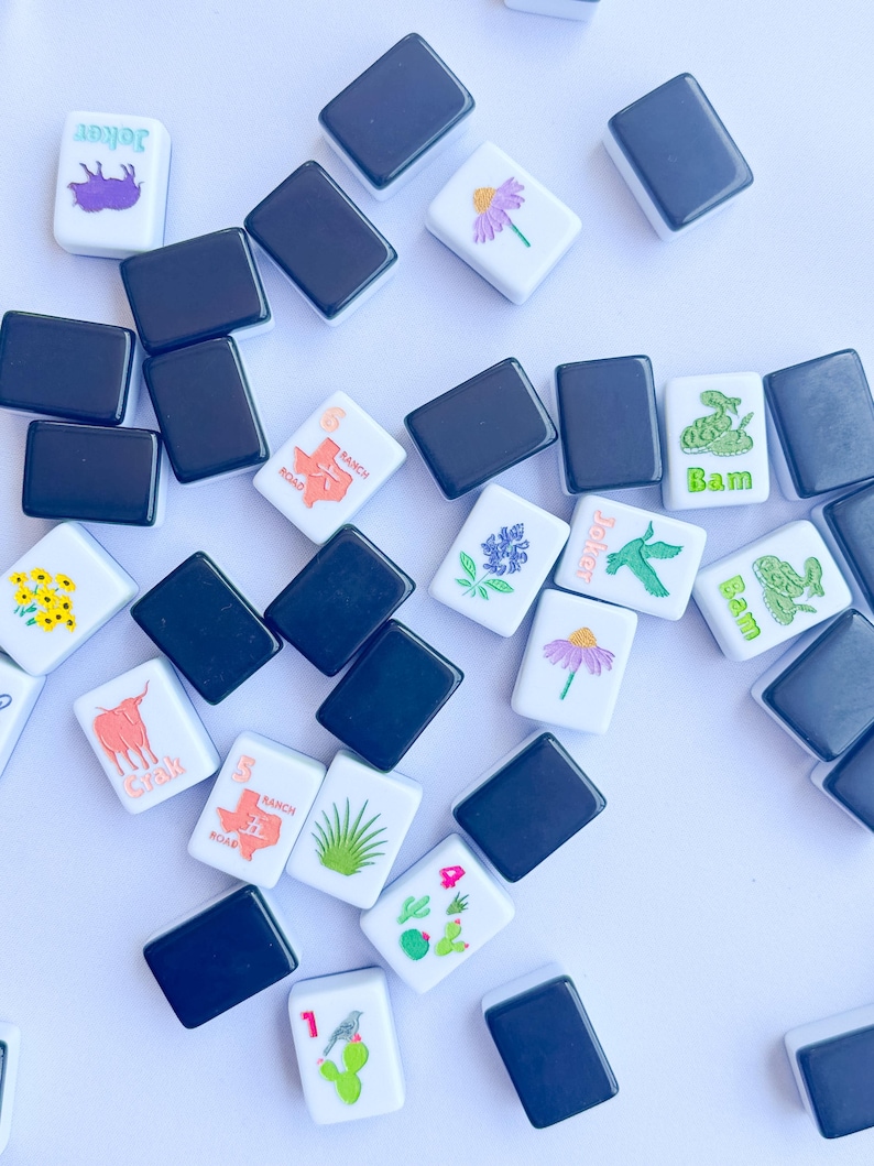 May include: A collection of white and black Mahjong tiles scattered on a white surface. The white tiles feature various colorful illustrations, including flowers, a longhorn, a cactus, and the text "Joker" and "Bam".