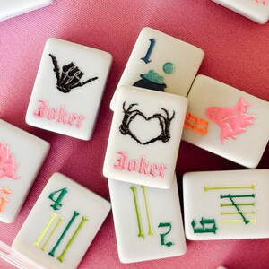 May include: A collection of white Mahjong tiles with various colorful symbols and numbers. Some tiles feature a black skeleton hand, a heart-shaped design, and the word "Joker" in pink. Other tiles display green and yellow lines and numbers.