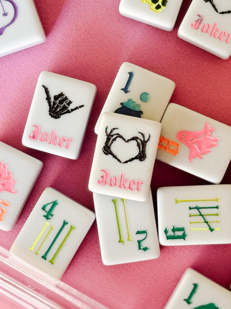 May include: A collection of white Mahjong tiles with various colorful symbols and numbers. Some tiles feature a black skeleton hand, a heart-shaped design, and the word "Joker" in pink. Other tiles display green and yellow lines and numbers.