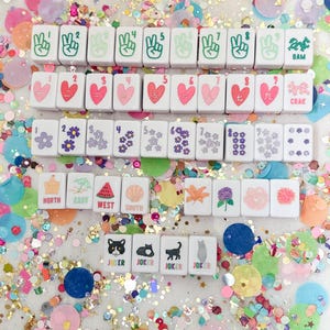 May include: A collection of white Mahjong tiles with various symbols and numbers, including peace signs, hearts, flowers, and directional indicators. The tiles are scattered amidst colorful confetti.