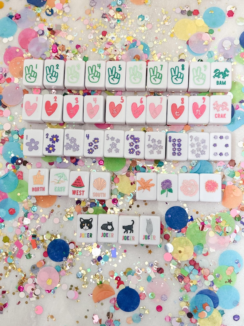 May include: A collection of white Mahjong tiles with various symbols and numbers, including peace signs, hearts, flowers, and directional indicators. The tiles are scattered amidst colorful confetti.