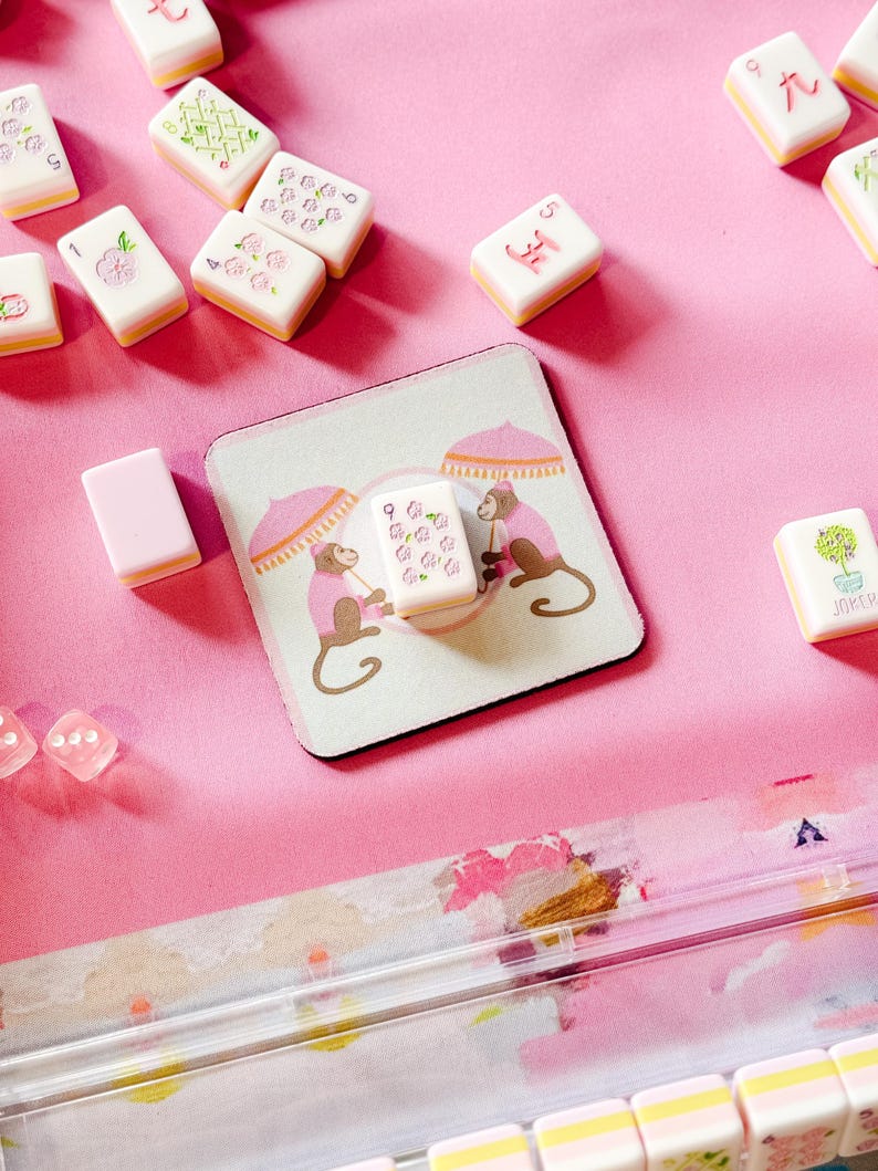 May include: A pink and white Mahjong set with tiles featuring floral and geometric designs. A matching coaster displays two monkeys under parasols. Dice and other game components are also visible, all set against a pink background.