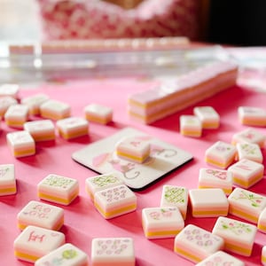 May include: A pink Mahjong set with white tiles featuring floral and character designs. The tiles have yellow and pink layers. The set includes a pink table surface and a coaster with a floral design.