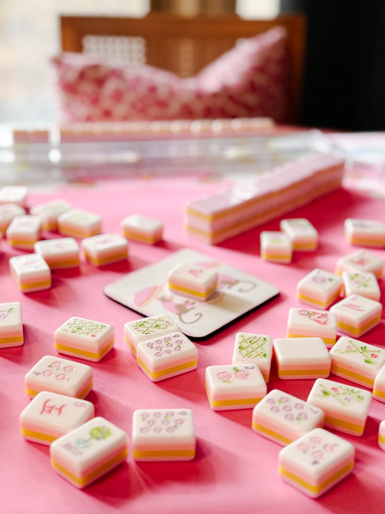 May include: A pink Mahjong set with white tiles featuring floral and character designs. The tiles have yellow and pink layers. The set includes a pink table surface and a coaster with a floral design.