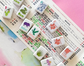 May include: A collection of white tiles with various illustrations and text, including a snake, a bird, and floral designs. The tiles are arranged on a patterned surface with printed text and numbers.
