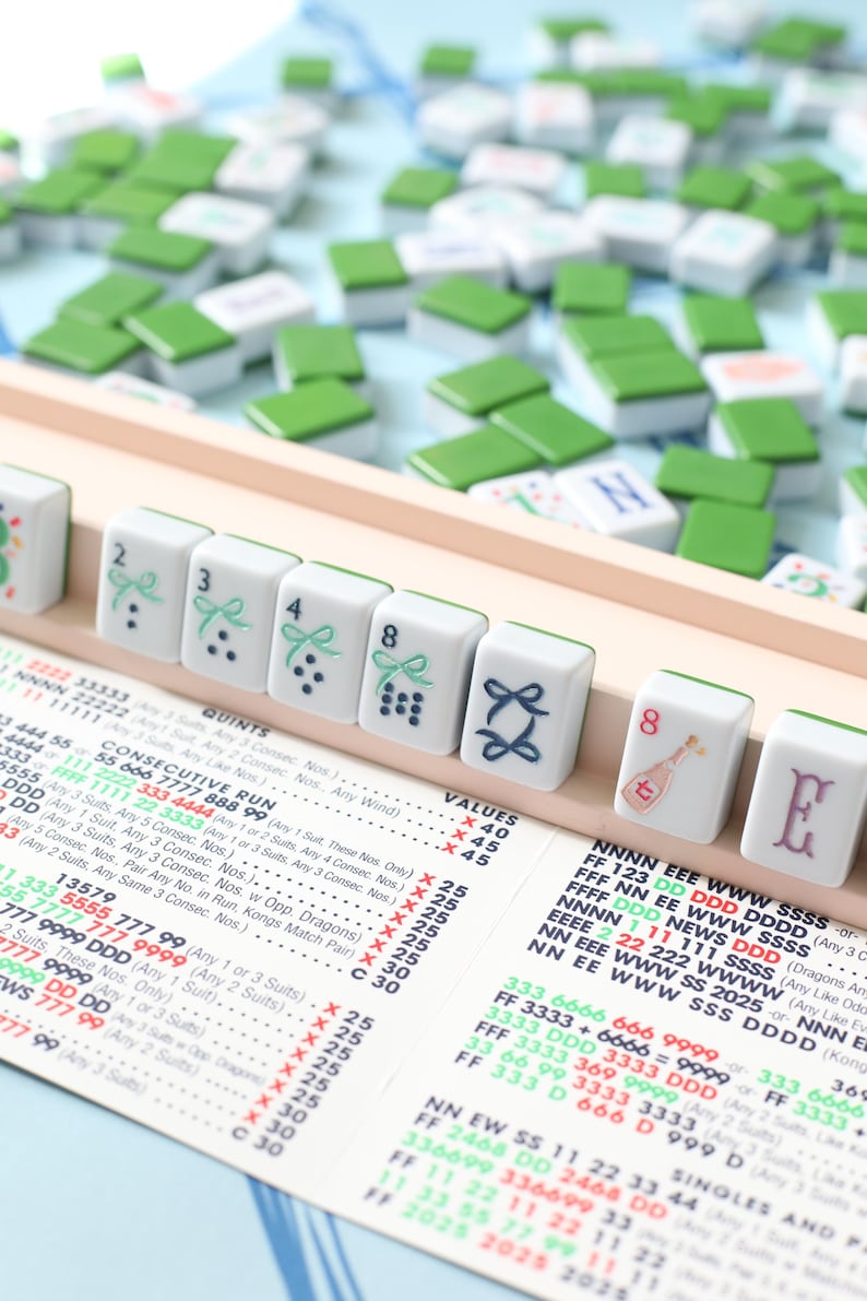May include: A close-up of a Mahjong game in progress, featuring white tiles with green tops. The tiles display various symbols and numbers, including the numbers 2, 3, 4, and 8, along with other unique designs. A game rules sheet is visible.