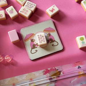 May include: A pink Mahjong set with tiles featuring floral and character designs. A coaster with monkey illustrations and a tile with the number nine is also visible. The set is arranged on a bright pink surface.
