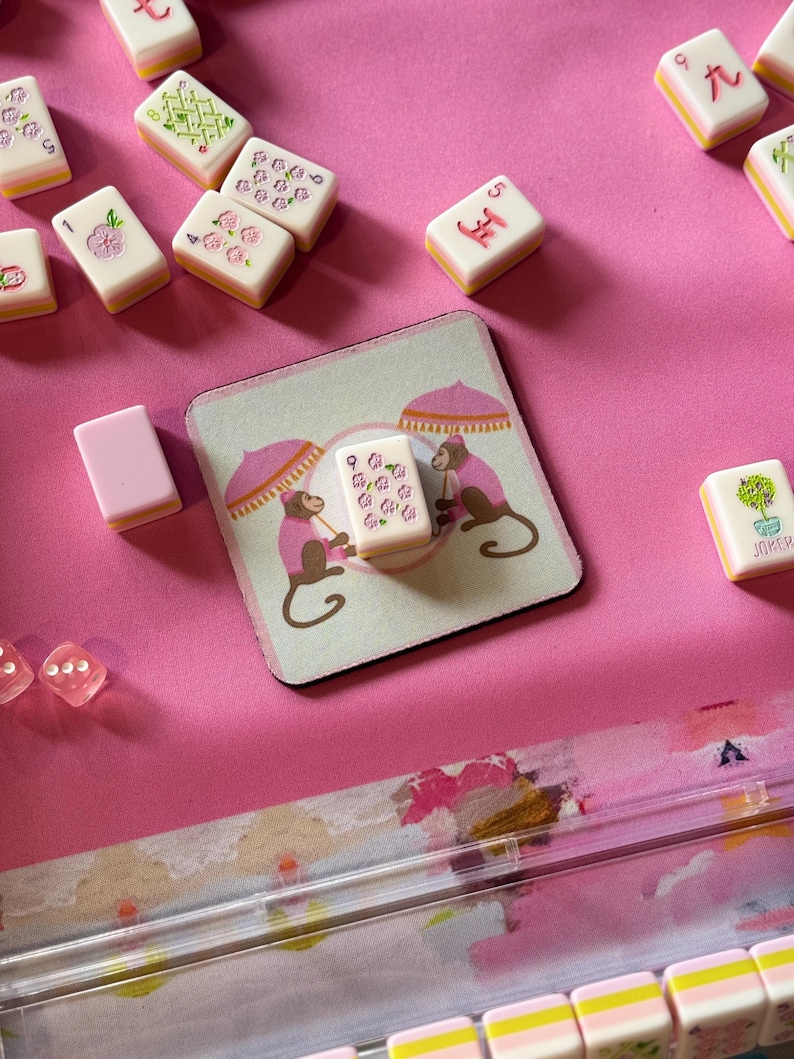 May include: A pink Mahjong set with tiles featuring floral and character designs. A coaster with monkey illustrations and a tile with the number nine is also visible. The set is arranged on a bright pink surface.