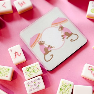 May include: A square coaster with a monkey design and several Mahjong tiles on a pink surface. The coaster features two monkeys under pink umbrellas. The Mahjong tiles are white with various symbols and numbers.