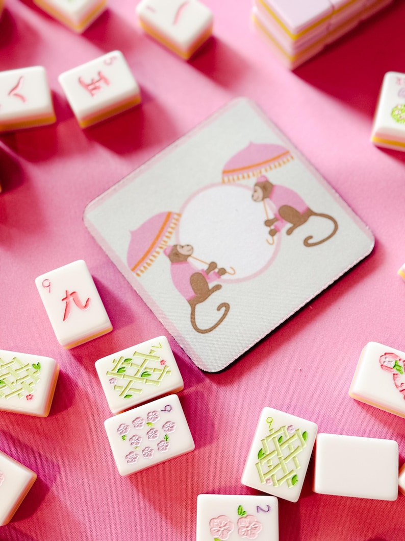 May include: A square coaster with a monkey design and several Mahjong tiles on a pink surface. The coaster features two monkeys under pink umbrellas. The Mahjong tiles are white with various symbols and numbers.