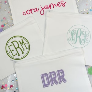 May include: Four white zippered pouches with different personalized designs. The top pouch has the text "cora james" in pink lettering. The pouch below has the initials "ERM" in green lettering inside a circle. The pouch below that has the initials "SR" in green lettering inside a circle. The bottom pouch has the initials "DRR" in purple lettering.