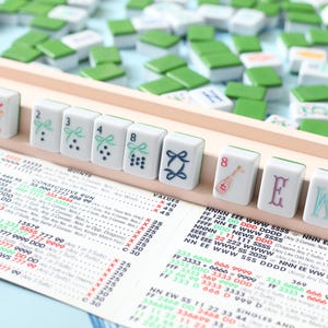 May include: A Mahjong set with white tiles featuring green and blue designs, numbers, and letters. The tiles are arranged on a wooden rack, with a game chart visible below and scattered tiles in the background.