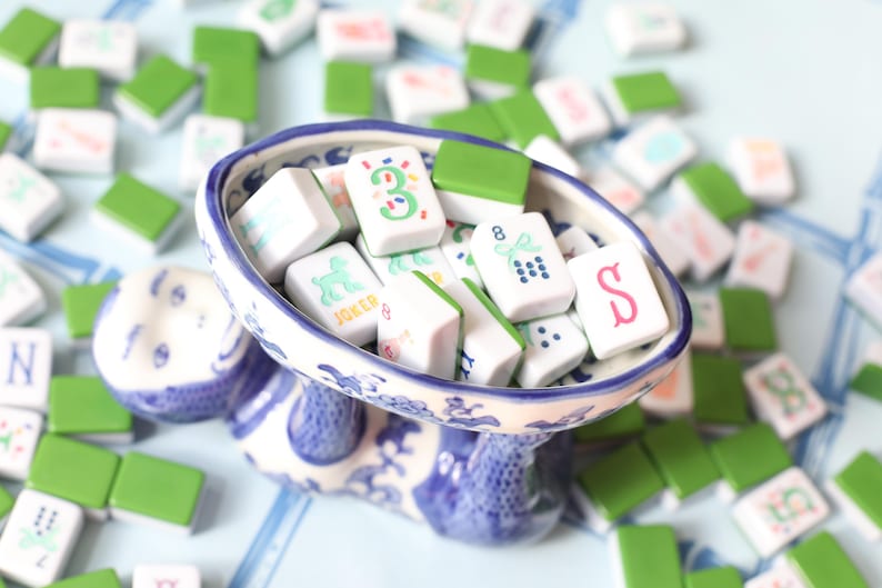 May include: A blue and white ceramic serving dish filled with Mahjong tiles. The tiles are white with green, red, and black symbols and numbers. Green tiles are scattered around the dish. The dish is supported by a decorative base.