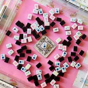 May include: A collection of Mahjong tiles scattered on a pink surface. The tiles are white with various symbols and numbers in blue, red, and green, alongside black tiles. A card with the text "MAHJOWEEN" is also present, suggesting a themed game.