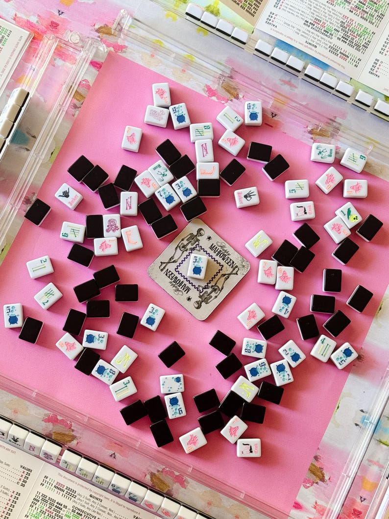 May include: A collection of Mahjong tiles scattered on a pink surface. The tiles are white with various symbols and numbers in blue, red, and green, alongside black tiles. A card with the text "MAHJOWEEN" is also present, suggesting a themed game.