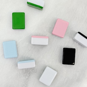 May include: An assortment of rectangular, glossy tiles in various colors, including green, pink, black, white, and blue. Some tiles have a two-tone design with white and a contrasting color. The tiles are scattered on a light-colored surface.