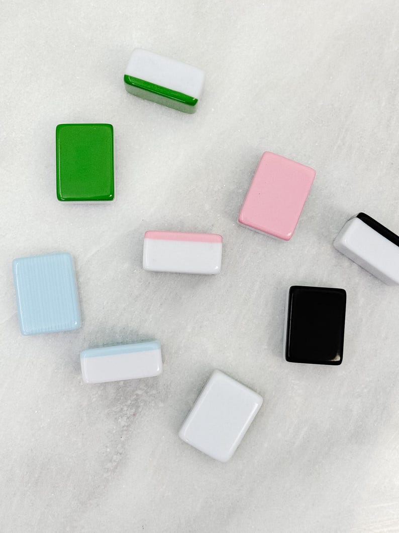 May include: An assortment of rectangular, glossy tiles in various colors, including green, pink, black, white, and blue. Some tiles have a two-tone design with white and a contrasting color. The tiles are scattered on a light-colored surface.