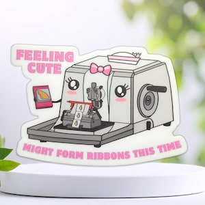 May include: A die-cut sticker featuring a cartoon sewing machine with a pink bow, large eyes, and rosy cheeks. The text reads "FEELING CUTE" and "MIGHT FORM RIBBONS THIS TIME" in pink. The machine holds a small pink frame with a yellow shape inside.