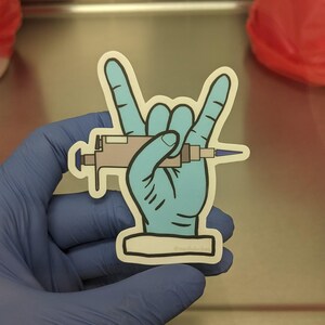 Peut inclure: Une main bleue avec une pipette fait le geste rock and roll. L'autocollant porte le texte "@medlabmemes" en bas.