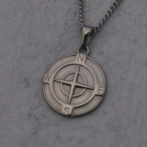 May include: A silver-colored compass pendant necklace. The pendant features a detailed compass rose design with the cardinal directions: North, South, East, and West. The necklace hangs on a matching silver chain, set against a gray background.