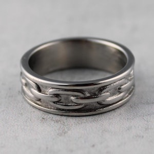 May include: A silver ring featuring a chain link pattern around the band. The ring is made of a shiny metal and is displayed on a light grey surface. The chain links are embossed into the metal.