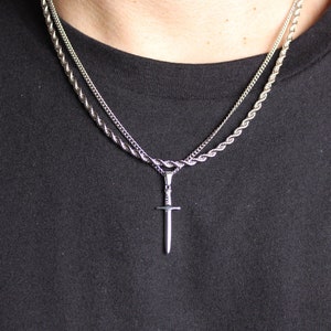 May include: A silver chain necklace with a silver sword pendant. The necklace is layered with a second, thinner chain.