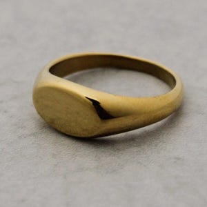 May include: A gold signet ring with a smooth, oval face. The ring is made of a shiny, yellow metal and sits on a light gray surface. The ring's design is simple and classic, suitable for everyday wear.