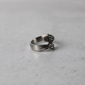 May include: A silver ring with two hands clasped together, forming a circle. The hands are detailed with intricate designs.