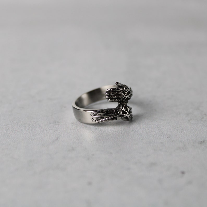 May include: A silver ring with two hands clasped together, forming a circle. The hands are detailed with intricate designs.