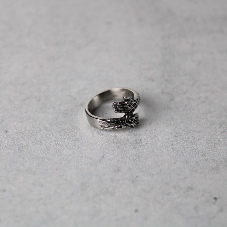 May include: A silver ring with two hands clasped together, forming a circle. The hands are detailed with intricate designs.