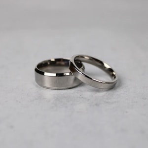 May include: Two silver wedding bands, one with a wider band and a beveled edge, the other with a thinner band.