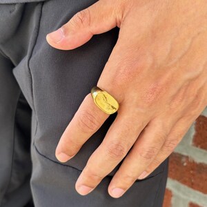 May include: A gold-colored signet ring with an oval face. The ring is worn on a finger, and the design on the face is visible. The ring appears to be made of metal and has a polished finish. The ring is a piece of jewelry.