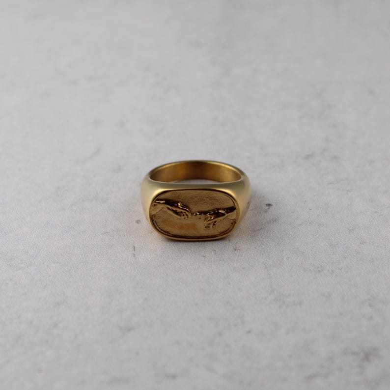 May include: A gold-colored signet ring with a rectangular face. The face of the ring features a detailed relief of two hands reaching towards each other, a design inspired by the famous artwork "The Creation of Adam."