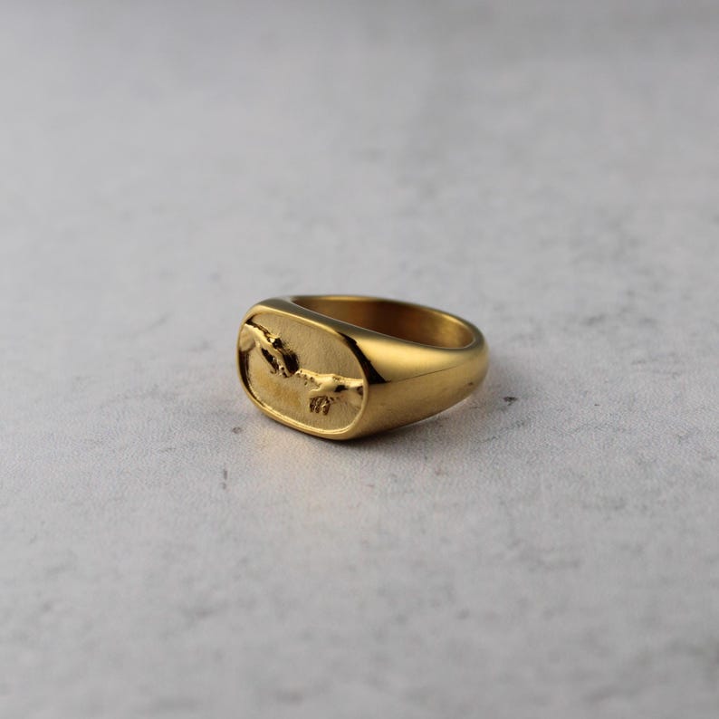 May include: A gold-colored signet ring with a rectangular face. The face of the ring features a detailed relief of two hands reaching towards each other. The ring has a polished, reflective surface and a thick band.