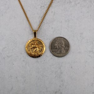 Puede incluir: Collar con colgante de San Crist&oacute;bal dorado. El colgante redondo presenta una imagen de San Crist&oacute;bal y el texto "SAINT CHRISTOPHER PROTECT US". La cadena del collar tambi&eacute;n es dorada. Una moneda est&aacute; al lado del colgante para comparar el tama&ntilde;o.