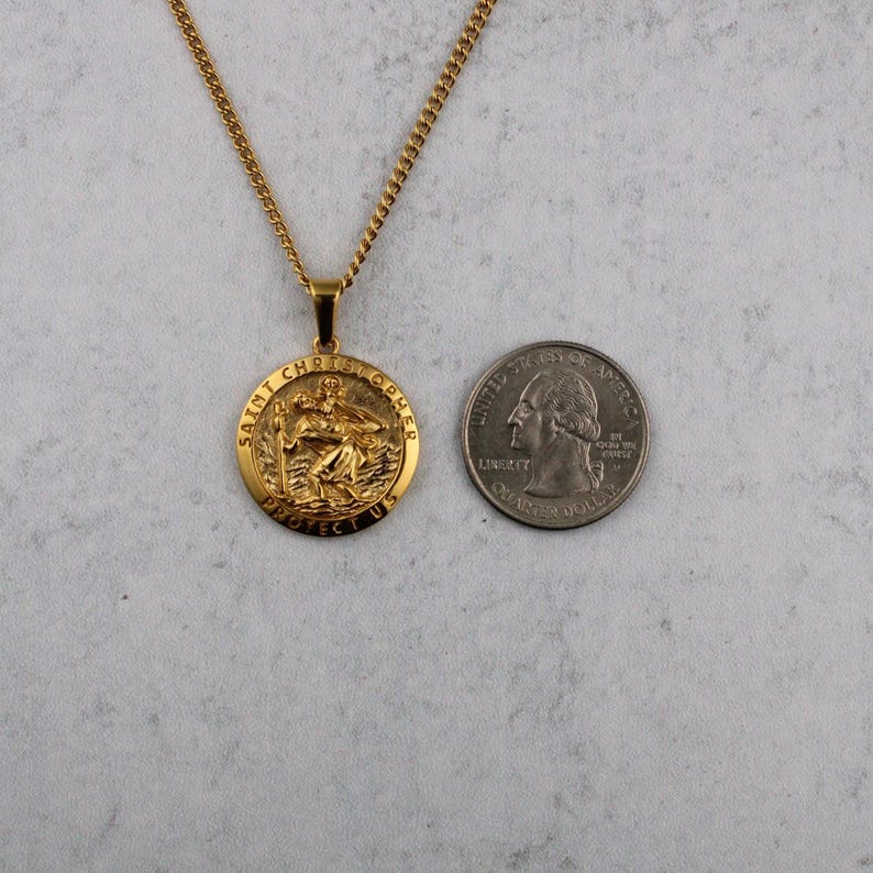 Puede incluir: Collar con colgante de San Crist&oacute;bal dorado. El colgante redondo presenta una imagen de San Crist&oacute;bal y el texto "SAINT CHRISTOPHER PROTECT US". La cadena del collar tambi&eacute;n es dorada. Una moneda est&aacute; al lado del colgante para comparar el tama&ntilde;o.