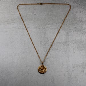 Puede incluir: Collar dorado con un colgante circular. El colgante presenta un dise&ntilde;o en relieve detallado. La cadena es de tipo eslab&oacute;n fino. El collar se muestra sobre una superficie gris claro.