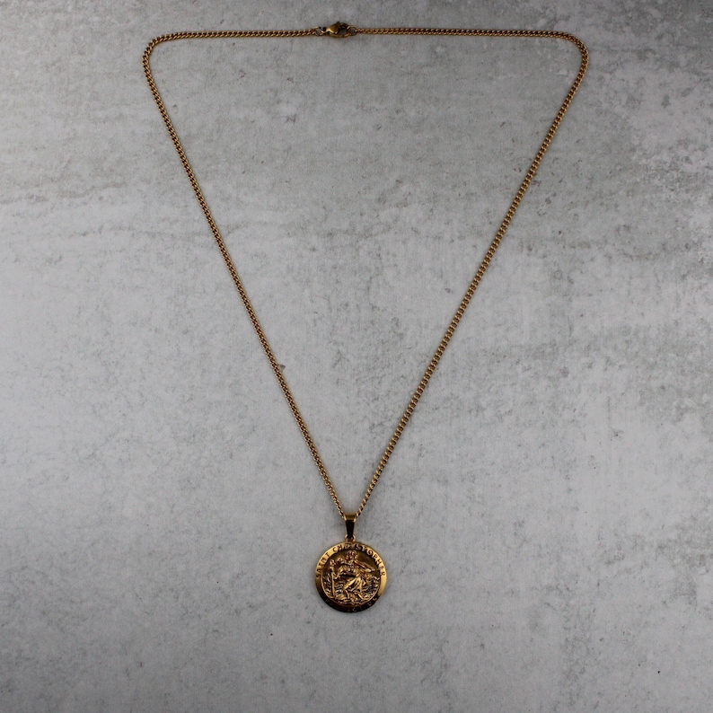 Puede incluir: Collar dorado con un colgante circular. El colgante presenta un dise&ntilde;o en relieve detallado. La cadena es de tipo eslab&oacute;n fino. El collar se muestra sobre una superficie gris claro.